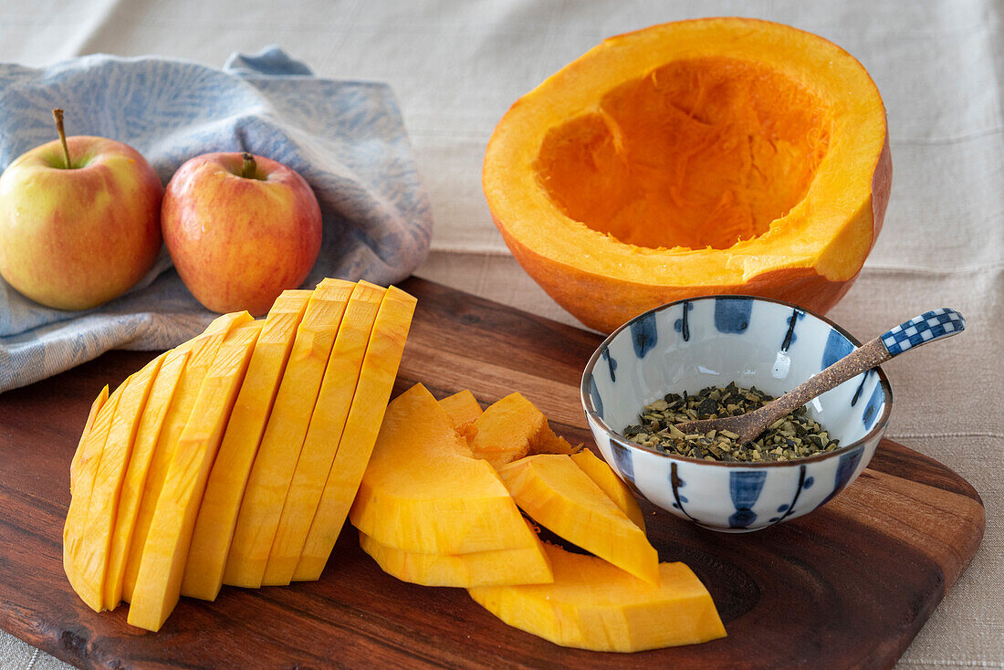 Sliced pumpkin and pumpkin seeds on wooden board, apples on tea towel