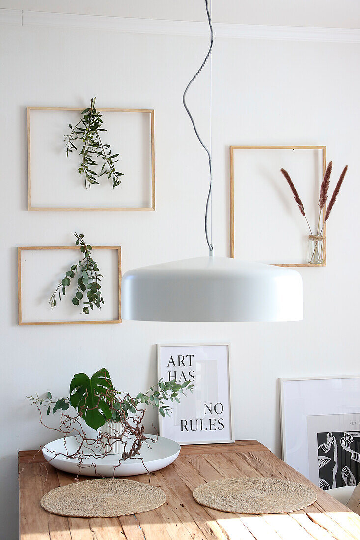 Dining area with hanging lamp, wooden table and plants in picture frames as wall decoration