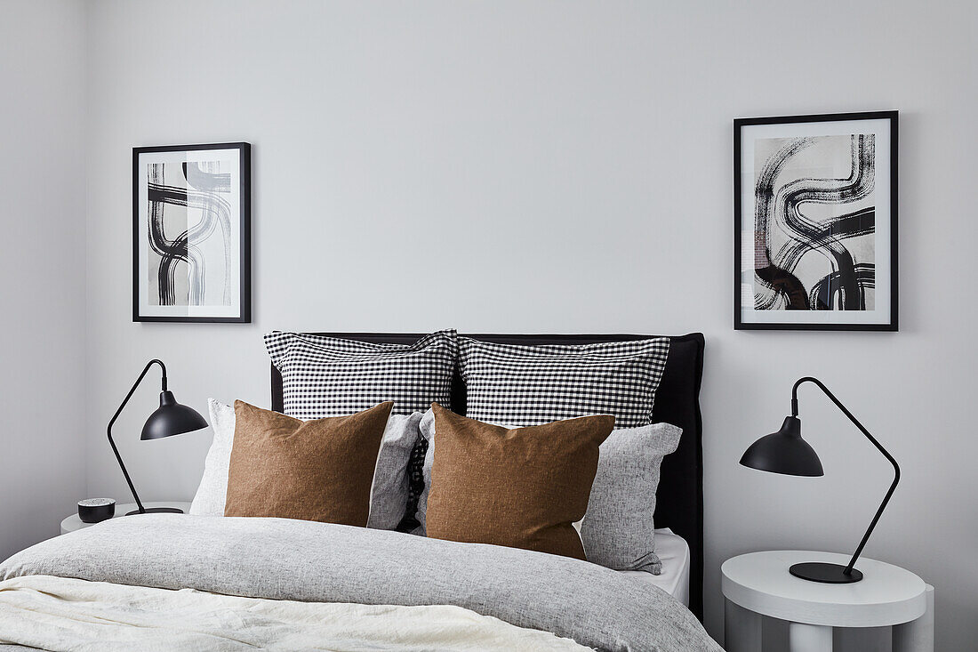 Modernes monochromes Schlafzimmer mit grauer und weißer Leinenbettwäsche und hellbraunen Kissen, abstrakten Bildern, weißen Beistelltischen und schwarzen Lampen