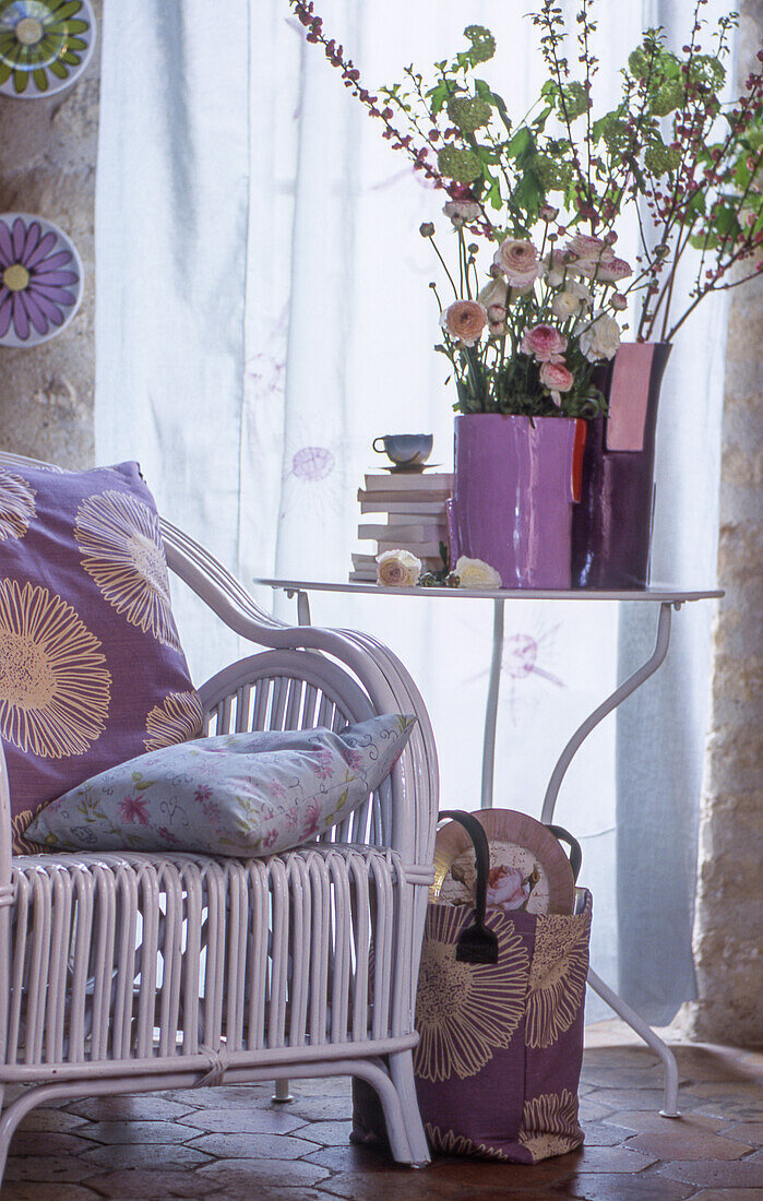 A white lacquered rattan chair with a cushion next to a side table with flowers and books