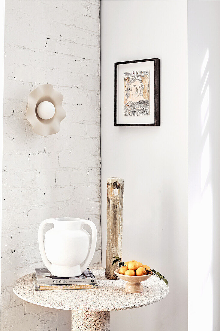 Round table with jug and fruit bowl with both portrait drawing and wall lamp above it