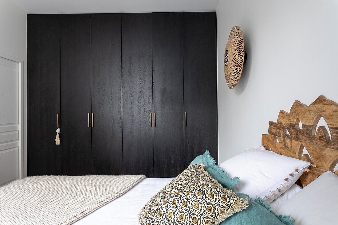 Doppelbett mit Kopfteil aus dekorativ geschnitzten Holzplatten und Kleiderschrank mit dunklen Fronten