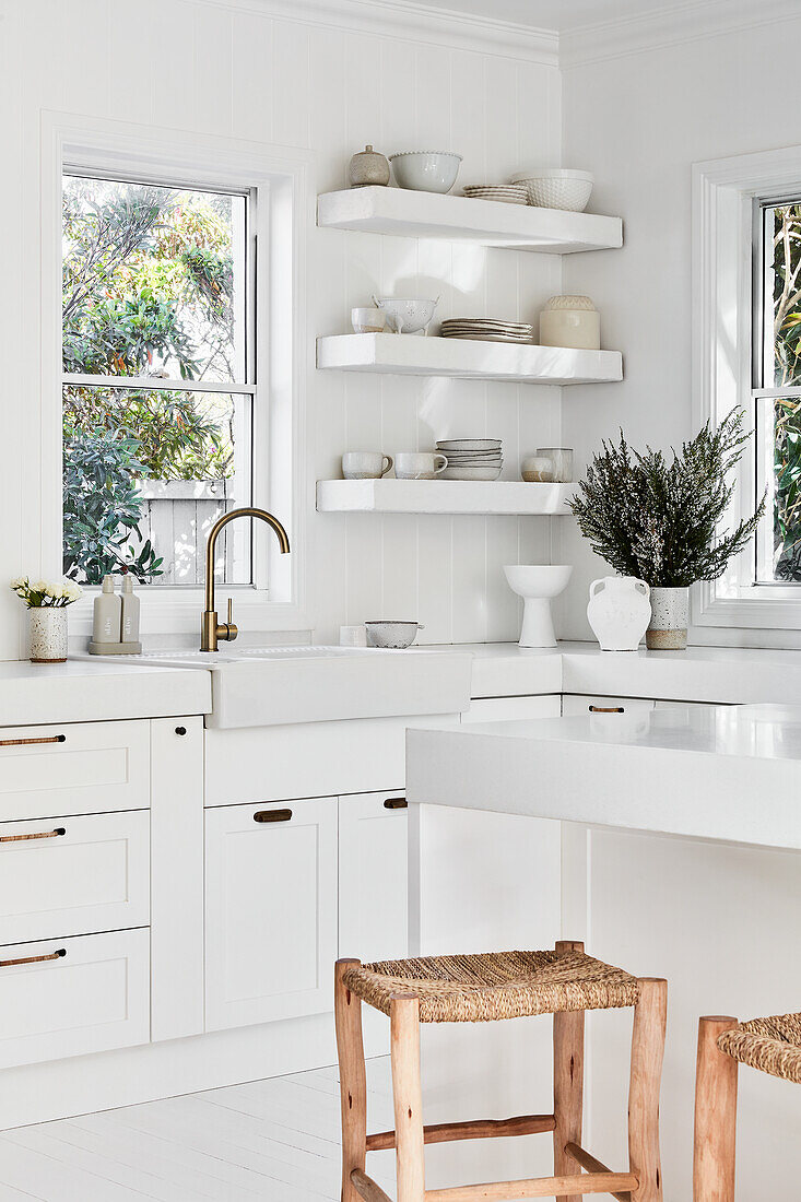 Weiße Küche mit Holzhockern um Kücheninsel, Porzellan-Spülbecken und offene Regale im Hintergrund