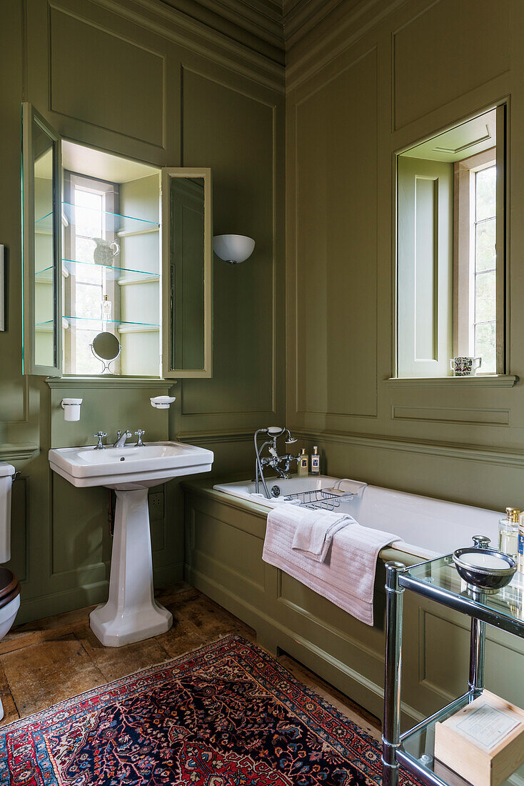 Olive green paneled bathroom with glass shelves in the window alcove and antique rug