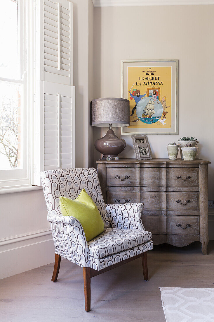 Upholstered armchair in front of window and a vintage chest of drawers in the corner of room