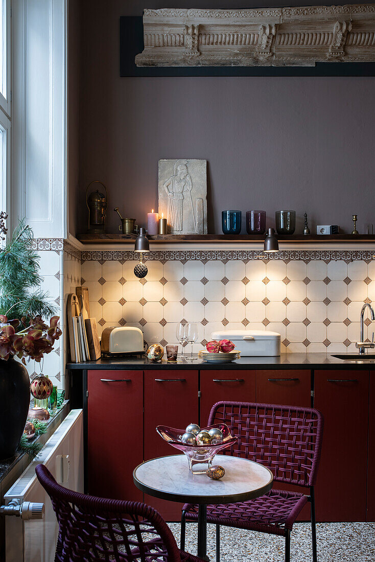 Dark red kitchenette, tiled backsplash and shelf above, small breakfast table in the foreground