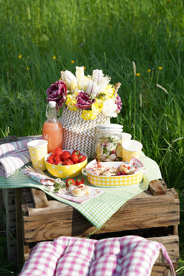 Holzkiste mit kariertem Tuch, Blumen. Leckereien und Obst auf Wiese im Frühling