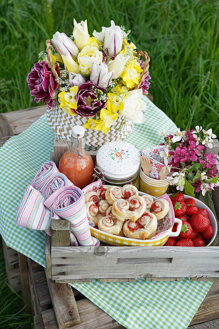 Picknickkorb mit Leckereien für ein Picknick auf Holzkiste mit kariertem Tuch und Blumenstrauß mit Tulpen