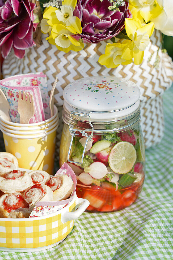 Vegetables in storage jar, wooden cutlery, mug, napkins and bouquet of flowers on picnic blanket