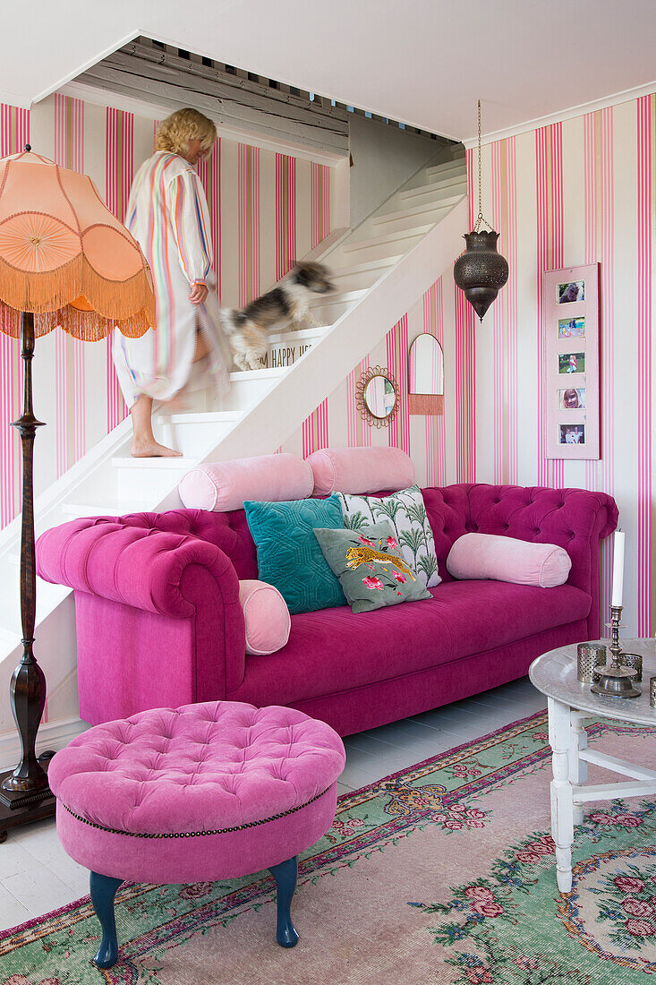 Pink sofa in lounge with pink striped wallpaper, white staircase with woman and dog