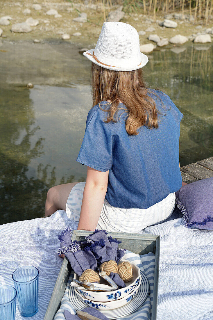 Woman sitting on wooden stick with view of the water, tray and crockery on blanket