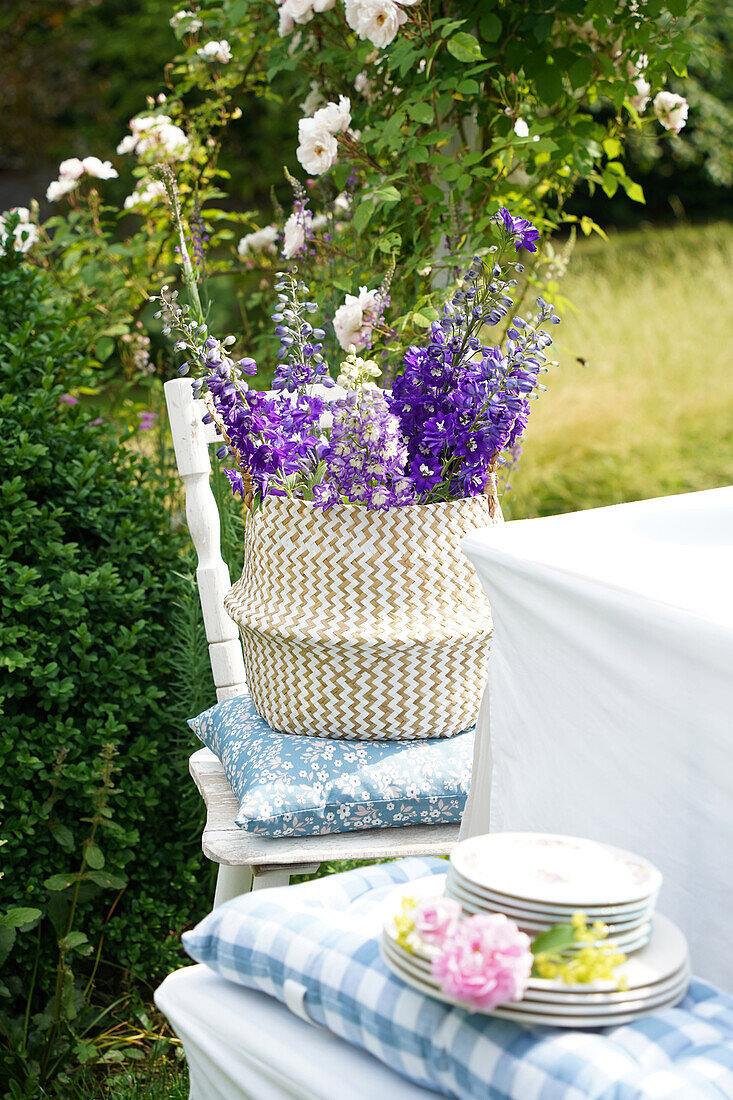 Delphinium bouquet in vase on garden chair with cushion, in the foreground pile of plates decorated with roses on bench