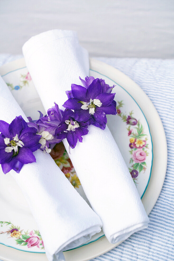 Serviettenringe aus violetten Ritterspornblüten (Delphinium) auf floralem Porzellanteller