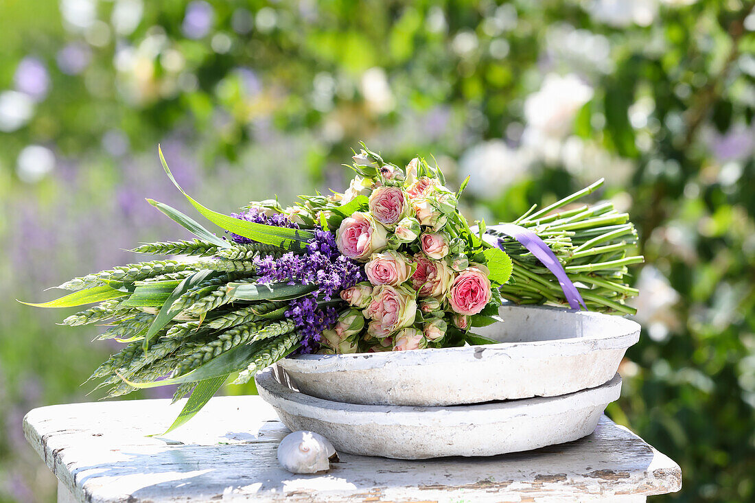 Blumenstrauß aus Rosen (Rosa), Lavendel (Lavandula) und Getreideähren auf Holzhocker im Garten