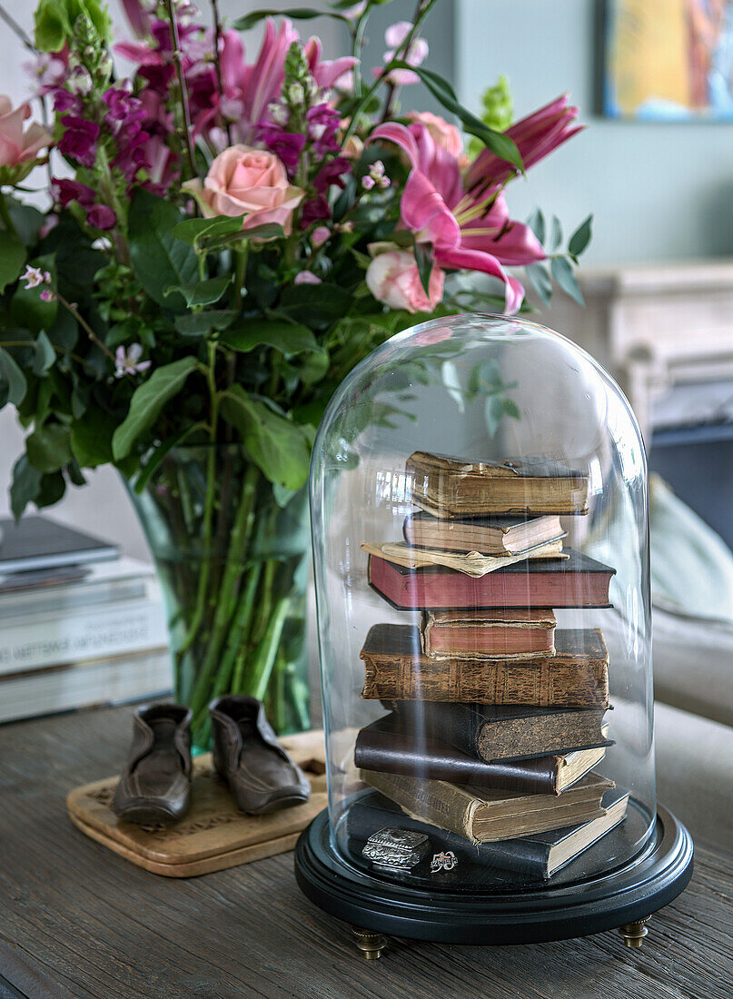 Stack of books under a glass bell on a wooden table next to a bouquet of roses and lilies