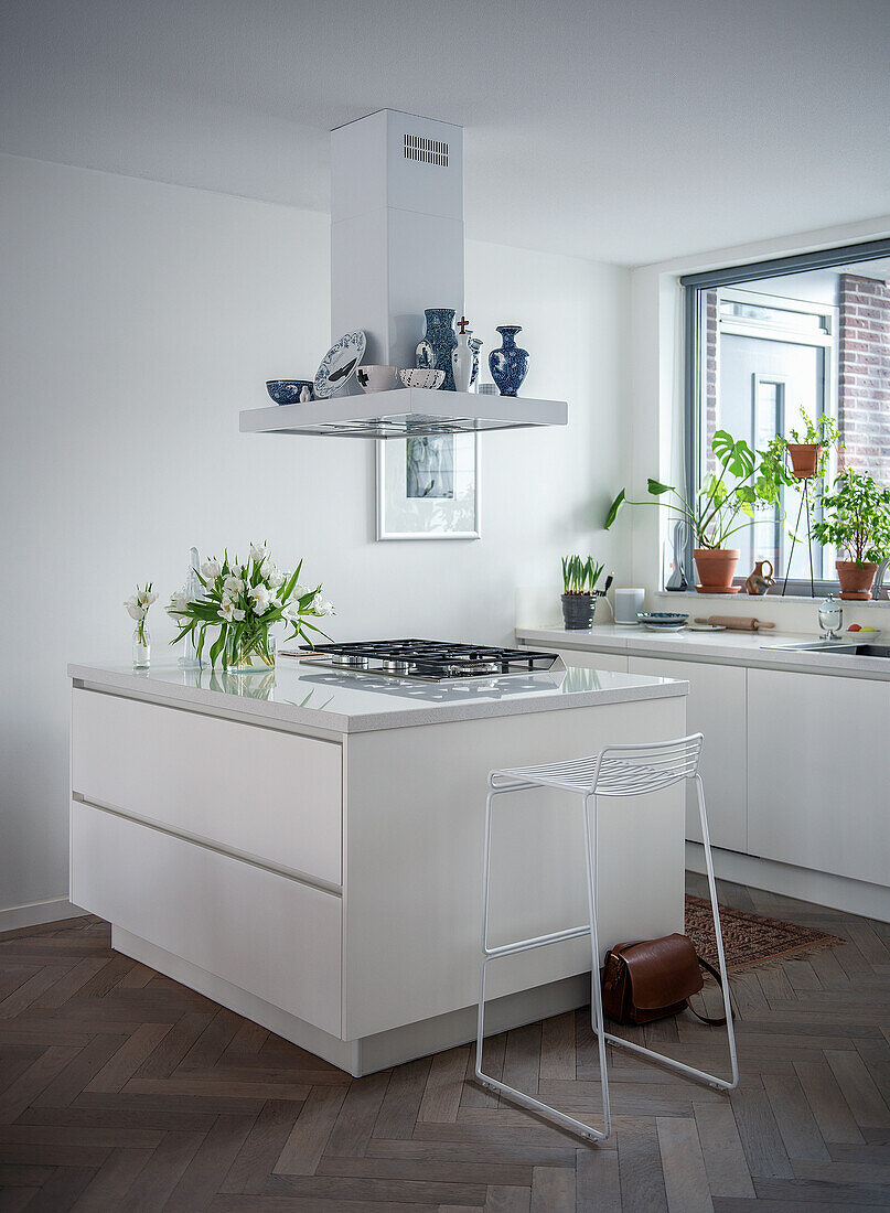 Modern white kitchen with cooking island and metal bar stool, decorated with plants and ceramic vases