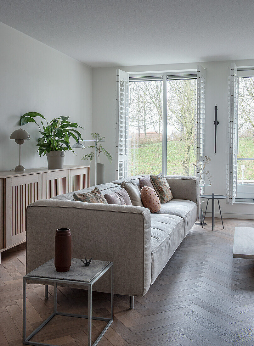 Modernes Wohnzimmer mit hellgrauem Sofa und Kissen, hölzerner Kommode und großen Fenstern