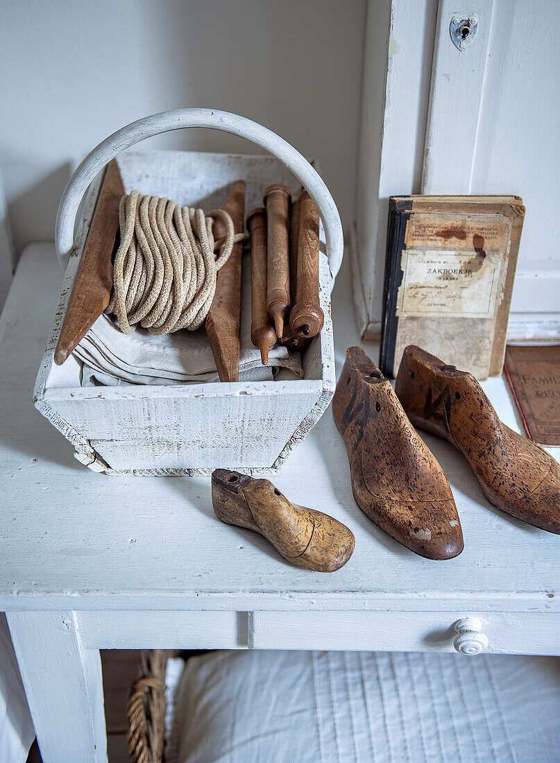 Rustikales Stillleben mit Holzkorb, Spule und altertümlichen Schuhformen auf weißem Nachttisch