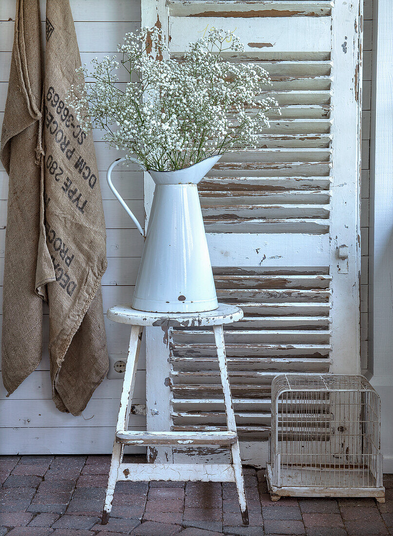 Large white jug with baby's-breath on a rustic stool in front of shutters