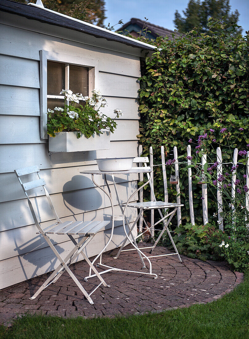 Weißer Bistro-Sitzbereich vor Gartenhaus mit Fensterblumenkasten