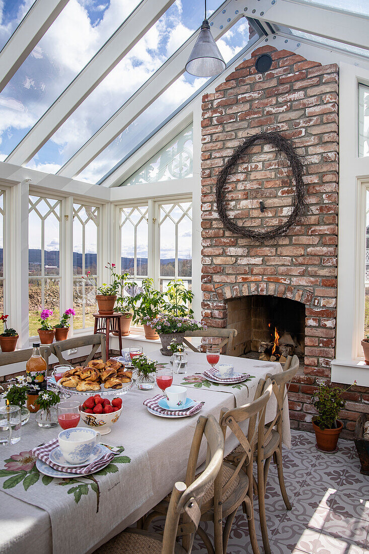 Laid table in a light-flooded conservatory with a brick fireplace and plants on a windowsill