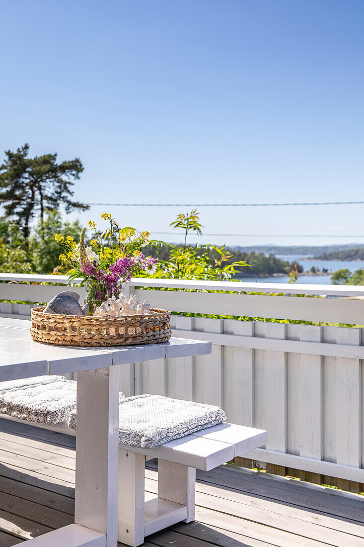 Geflochtenes Tablett mit Sommerblumenstrauß auf Holztisch auf Balkon mit Seeblick