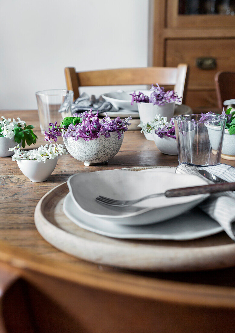 Gedeckter Holztisch mit weißen Tellern und lilafarbenen Blumenarrangements