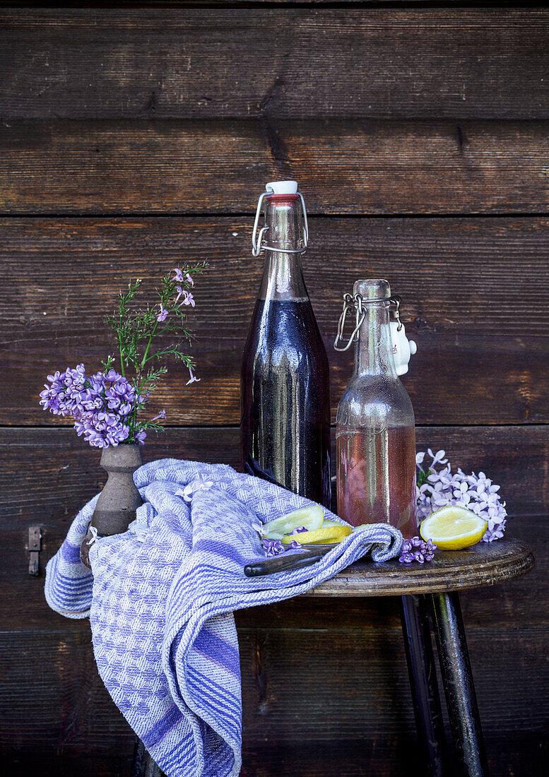 Glasflaschen mit Sirup, Zitronenscheiben und Lavendelblüten auf rustikalem Holzhocker