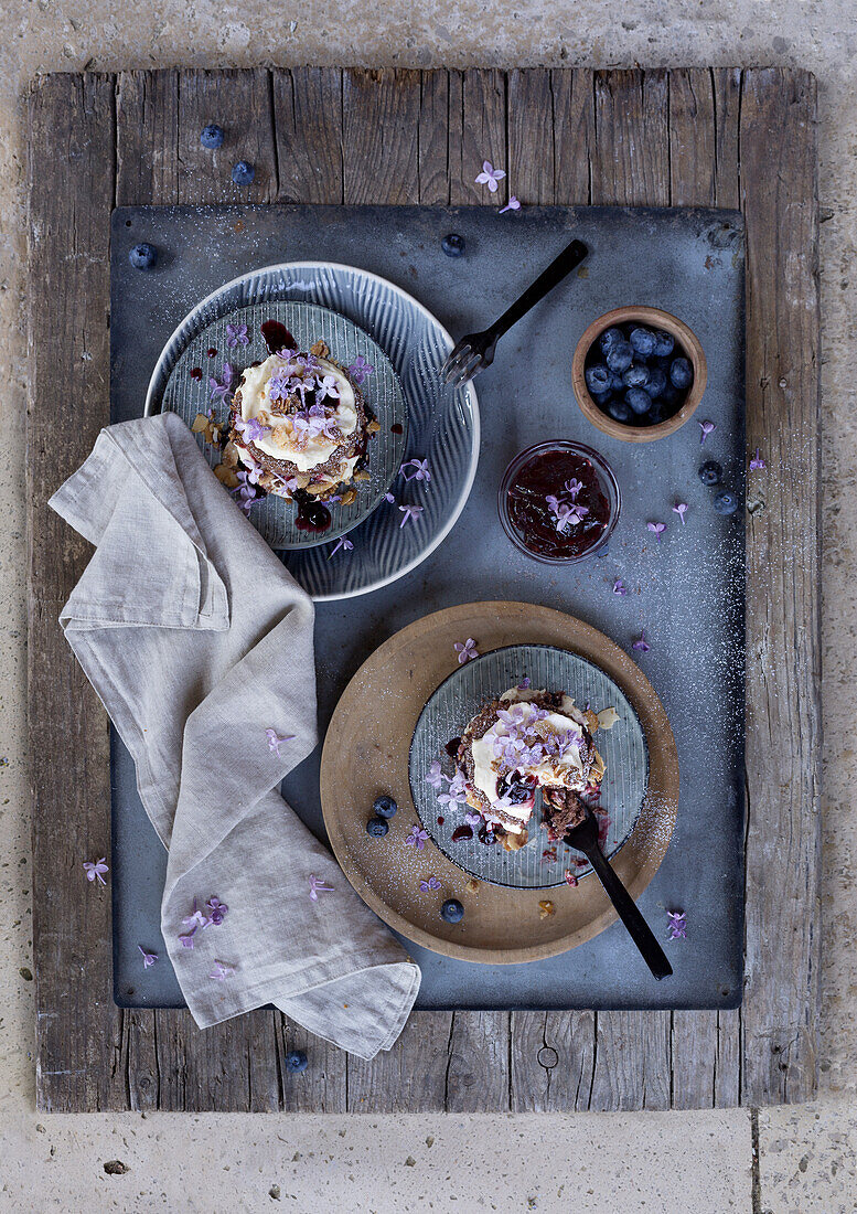 Frühstückstablett mit Pancakes, Heidelbeeren und essbaren Blüten auf rustikalem Holztisch