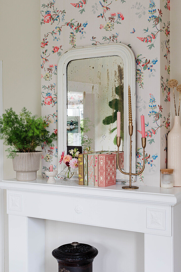 Decorative objects and wall mirror in front of floral wallpaper above white mantelpiece