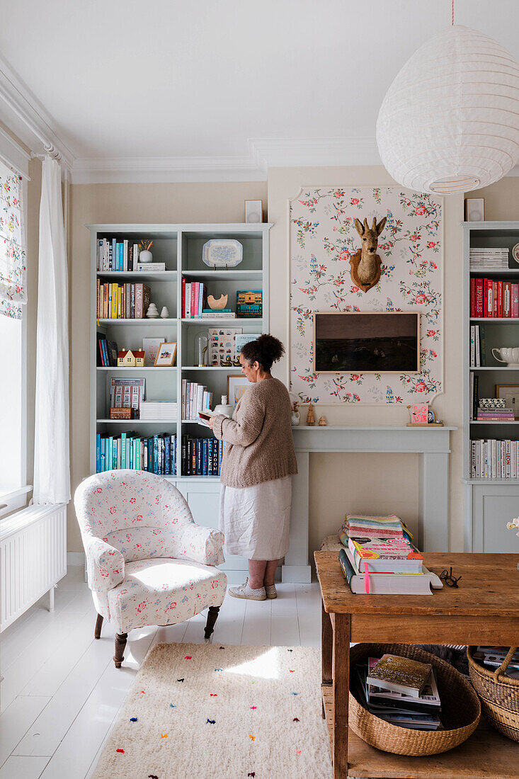 Person in front of bookshelf in living room with floral wallpapered wall