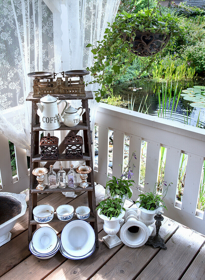 Wooden ladder shelf with vintage crockery on veranda overlooking pond