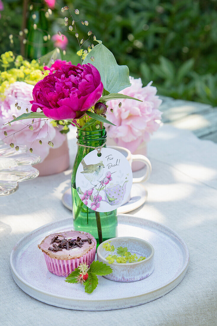 Tischdeko mit rosa Pfingstrose (Paeonia) in Glasflasche und Muffin auf Tablett