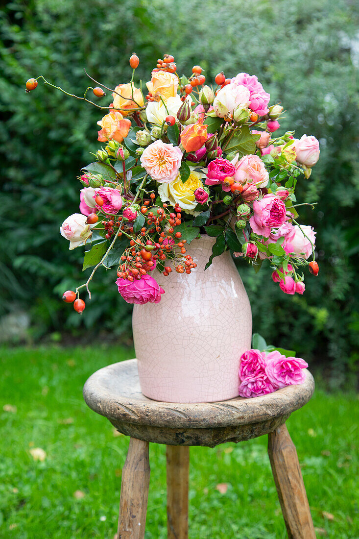 Rosenstrauß mit Hagebutten in rosa Vase auf Holzhocker im Garten