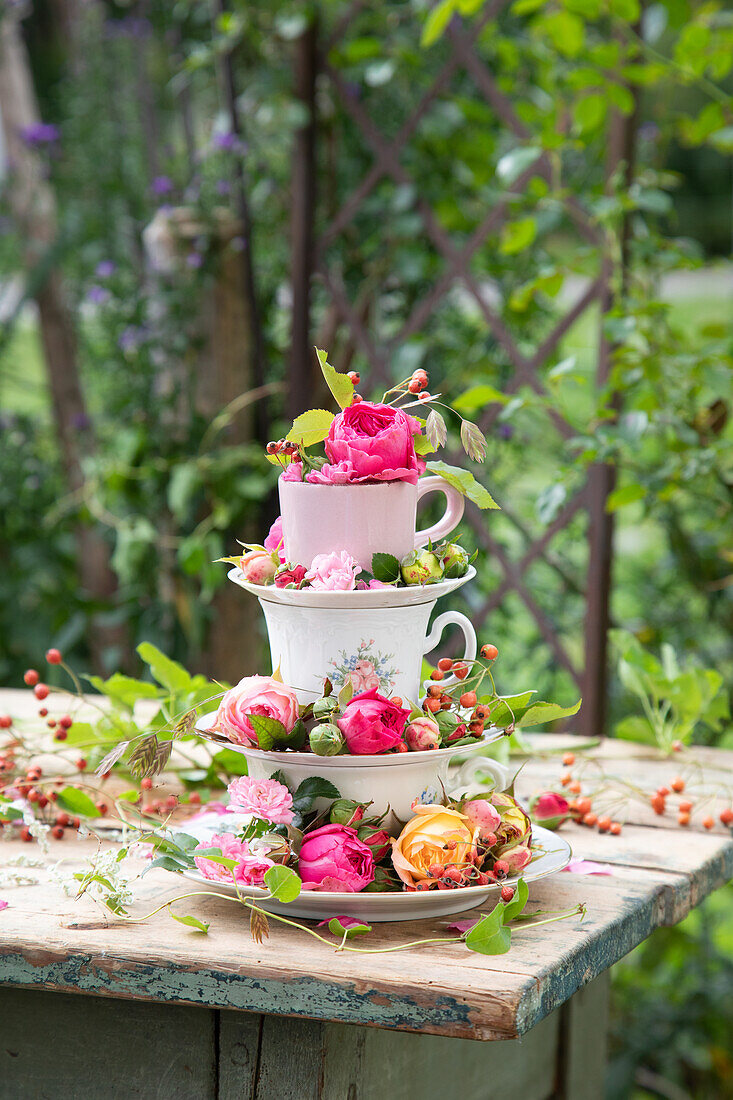 Stapeltassen mit Rosenblüten und Hagebutten auf rustikalem Holztisch im Garten