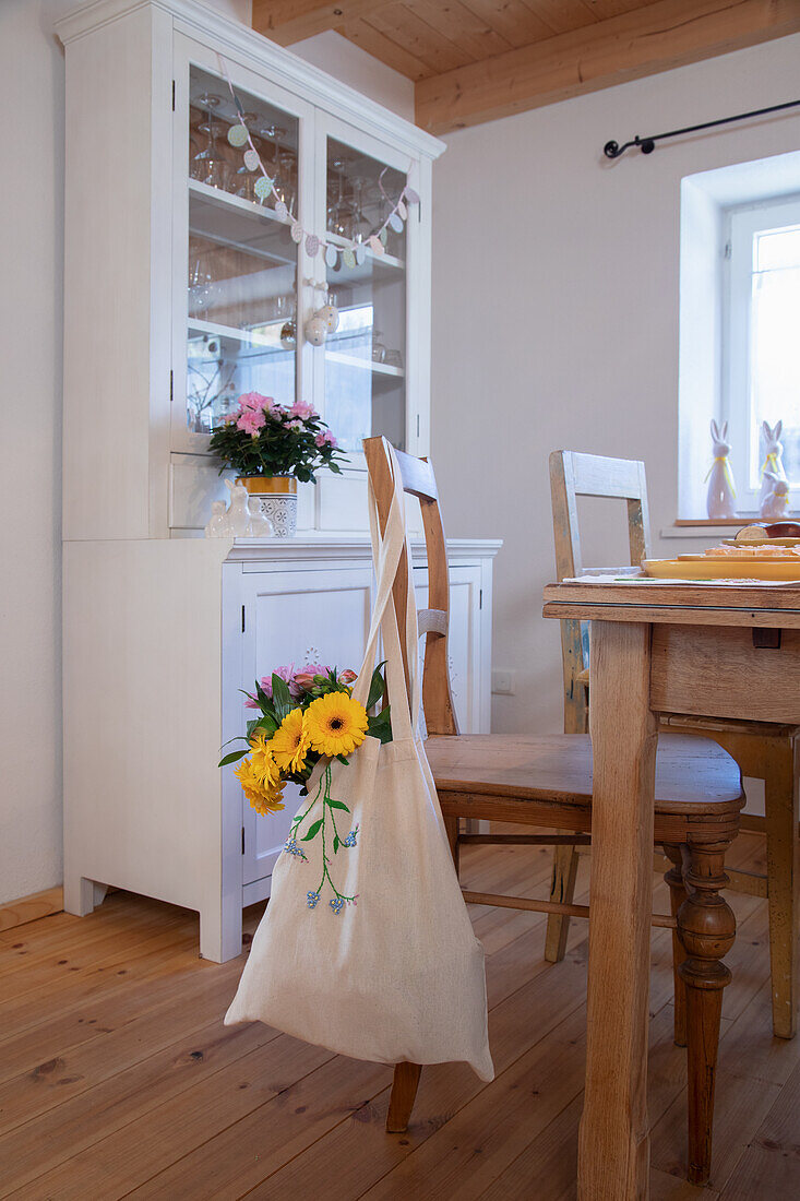 Esszimmer mit weißem Vitrinenschrank, Stofftasche hängt mit gestickten Frühlingsblumen am Stuhl