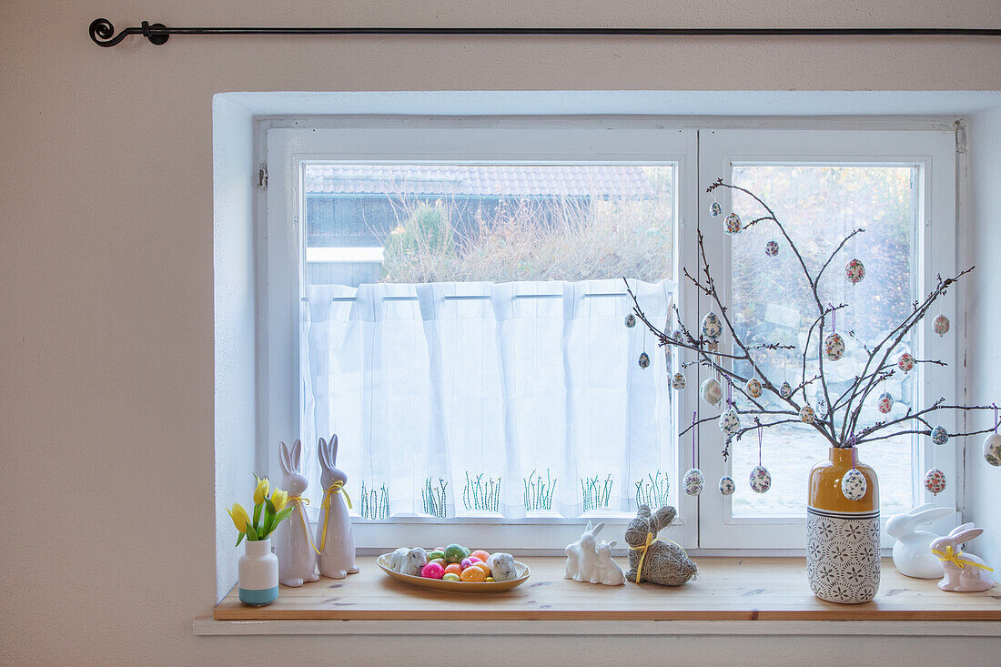 Osterdekoration auf Fensterbank mit bemalten Eiern, Hasenfiguren und Osterstrauß