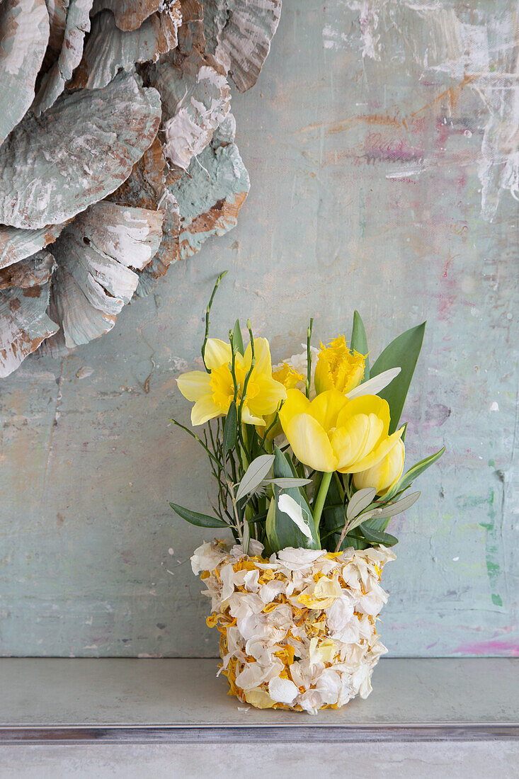 Mit Blütenblättern verzierter Blumentopf mit gelben Frühlingsblumen vor rustikalem Hintergrund