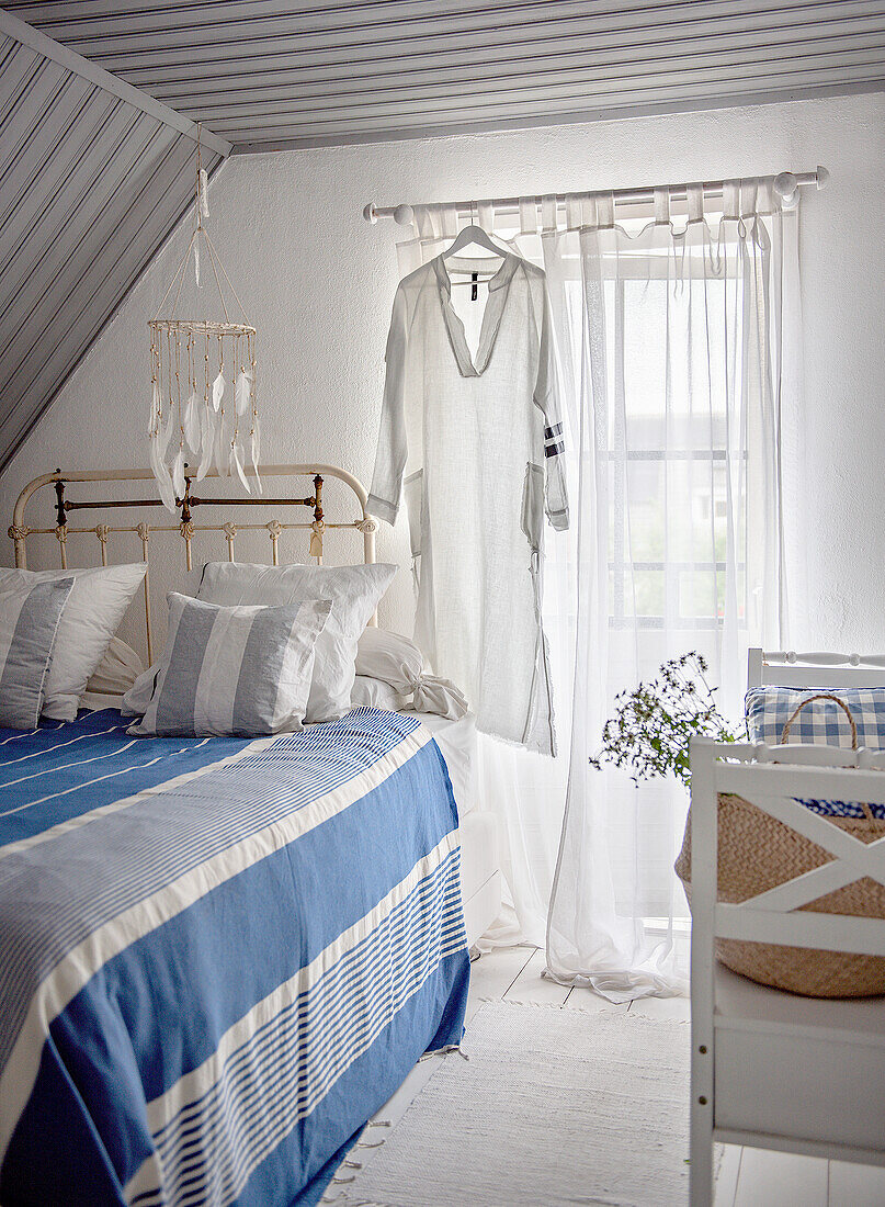 Schlafzimmer im Dachgeschoss mit blau-weiß gestreifter Bettwäsche und hängendem Kleid vor einem Fenster