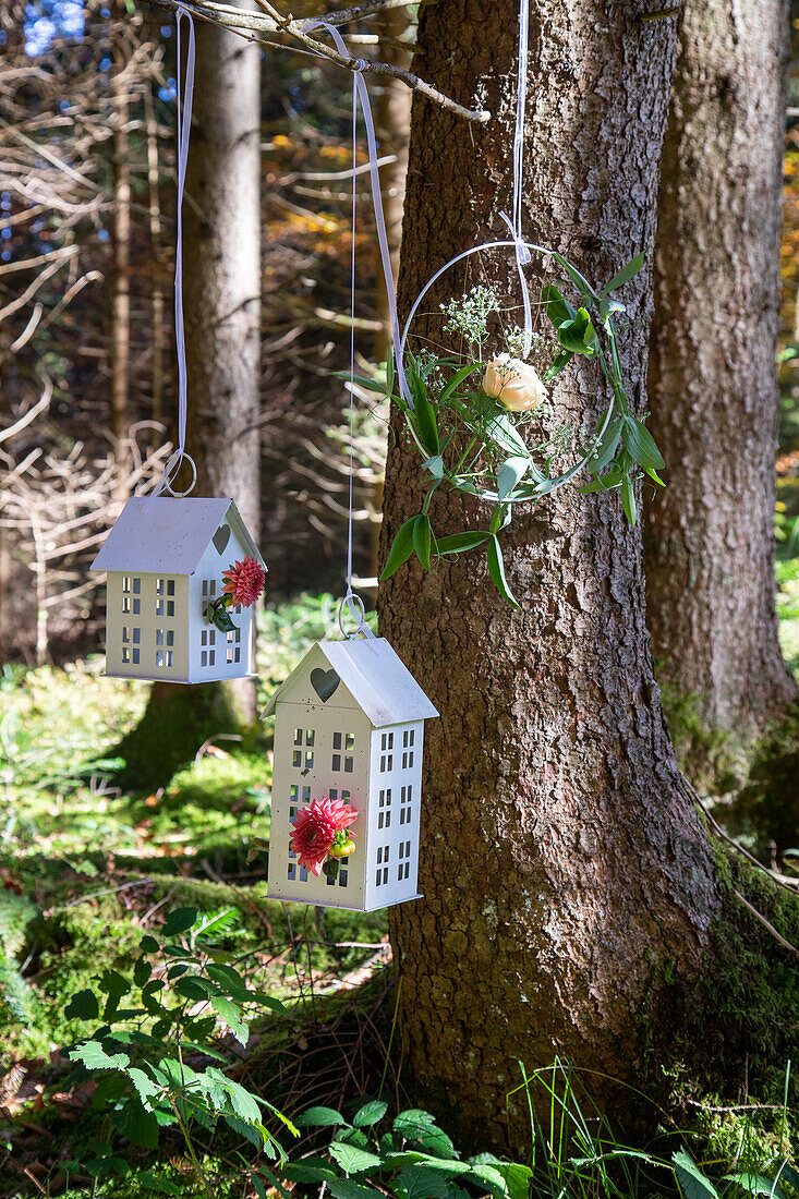 Laternen in Häuschenform und Blumenkranz hängen an Baum im Wald