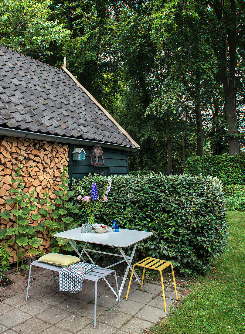 Kleiner Sitzbereich mit Metalltisch und gelbem Hocker neben Holzstapel im Garten