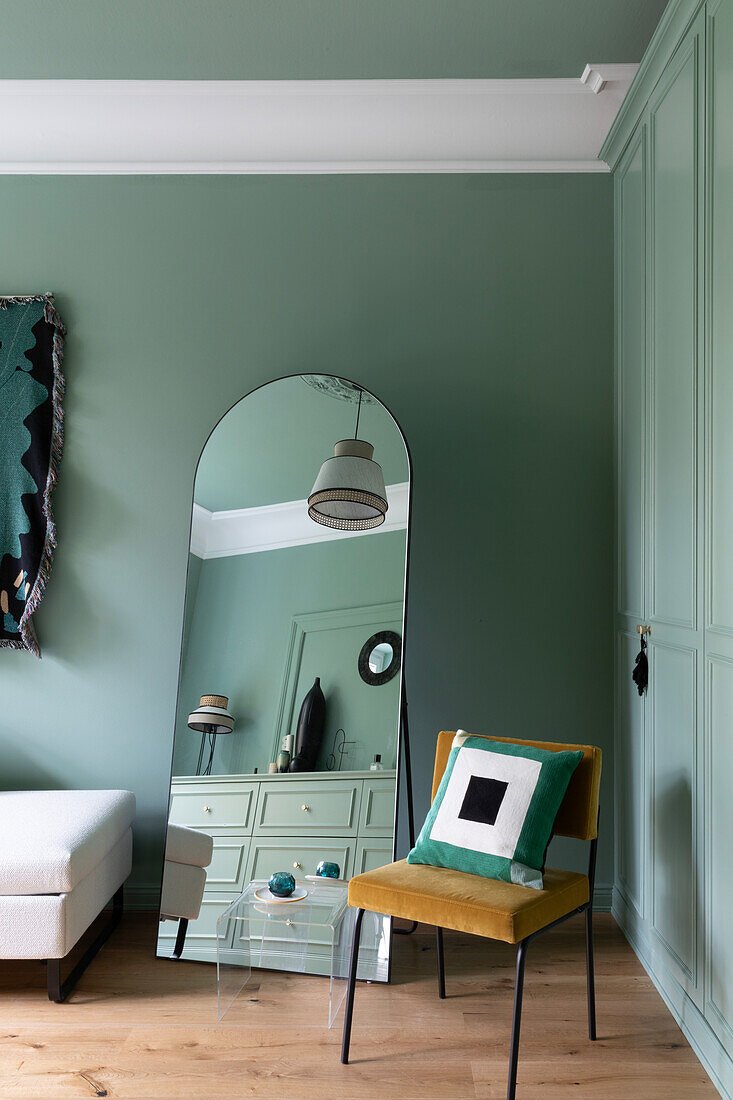 Bedroom with wardrobe and walls in pastel green, full-length mirror and chair with cushions