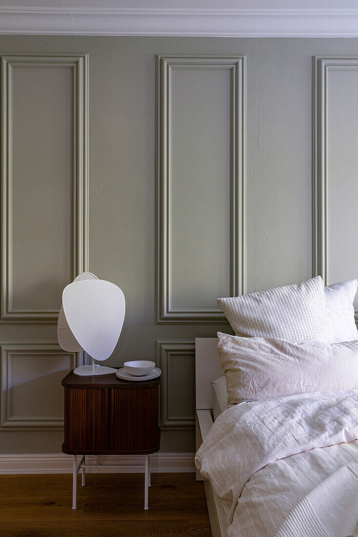 Wooden bedside table with table lamp, wall panelling in pastel green and bed with cushions and blanket