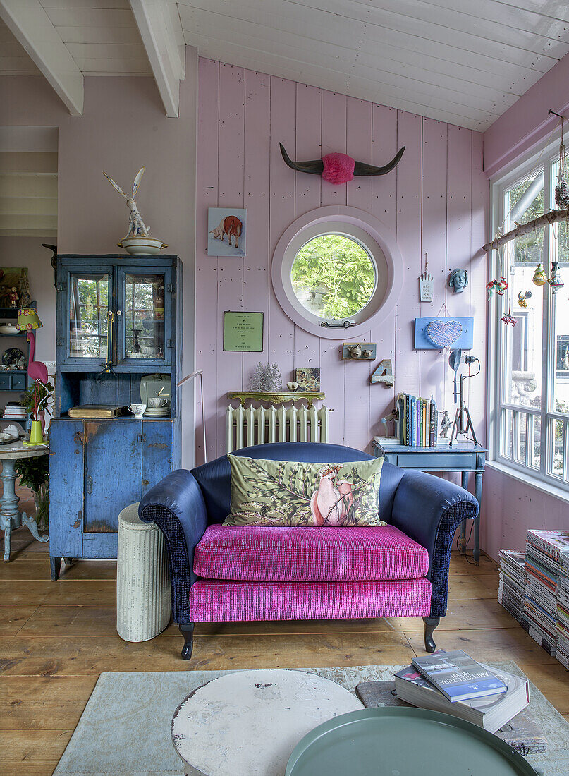 Wohnzimmer mit pink-blauem Sofa, Vitrine und runder Fensteröffnung