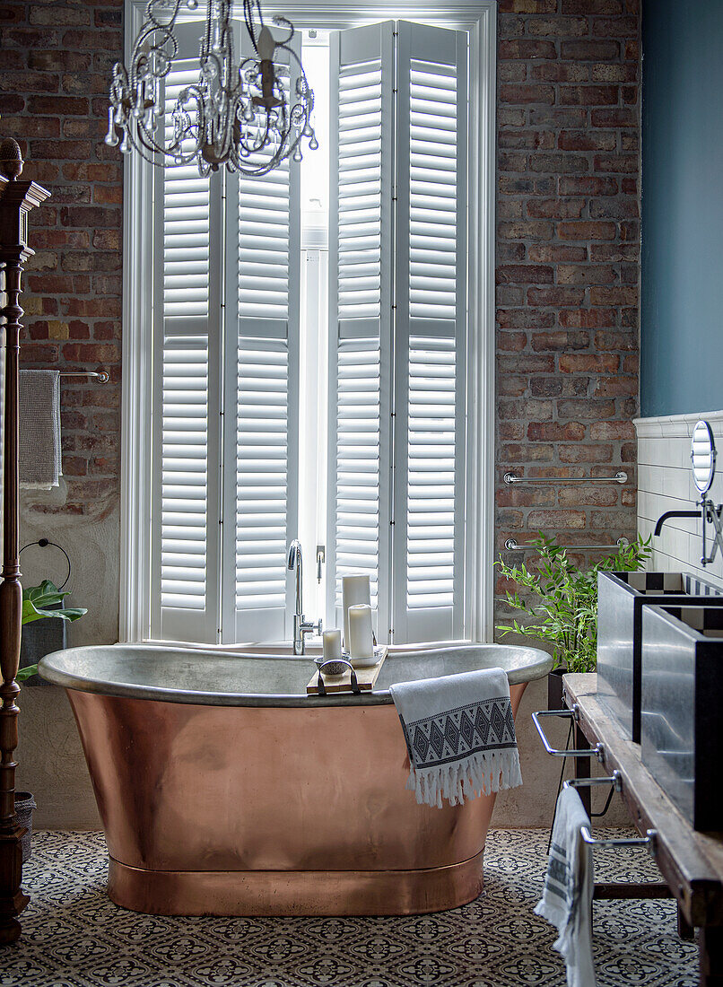 Free-standing copper bathtub in front of window with shutters and brick wall in bathroom