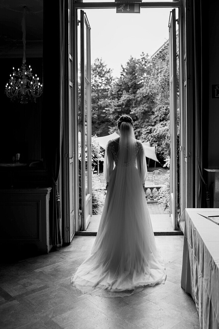 Bride in a long dress stands in front of an open door with a view of the garden