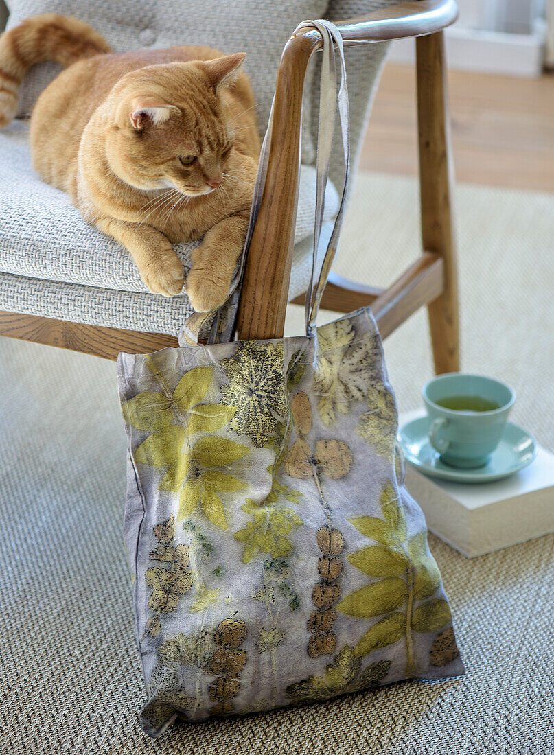 Stofftasche mit Blättermuster und Katze auf grauem Sessel