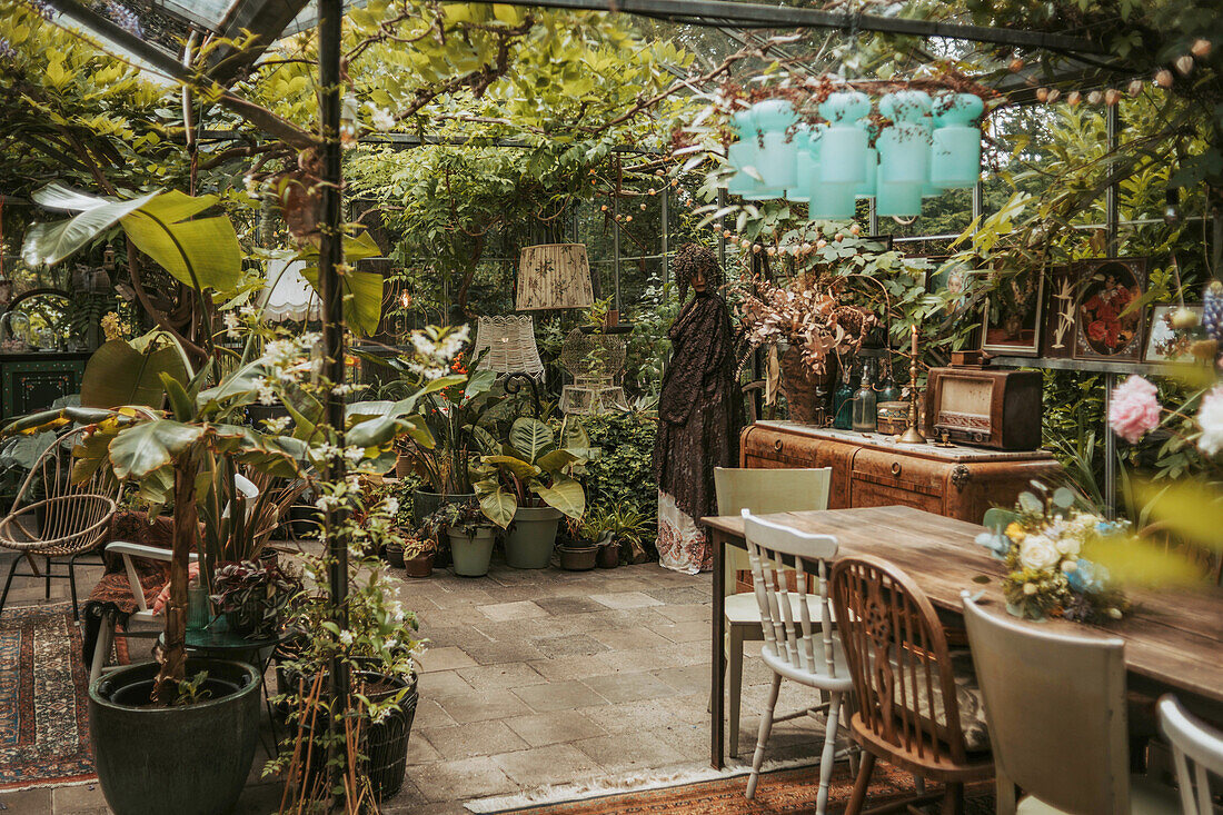 Gewächshaus mit üppigen Grünpflanzen, Holztisch mit Stühlen und Vintage-Deko