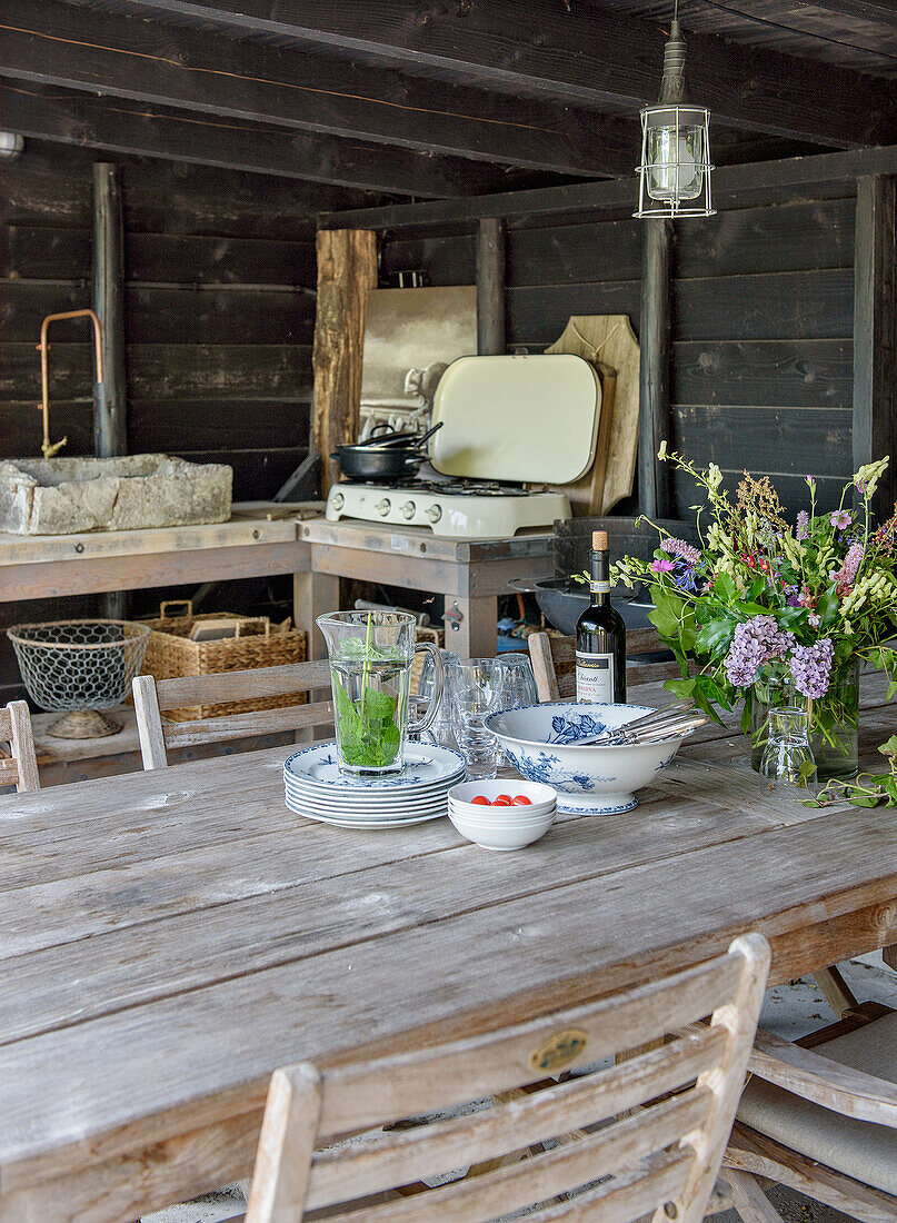 Set wooden table in a rustic gazebo with a bouquet of flowers and outdoor kitchen in the background