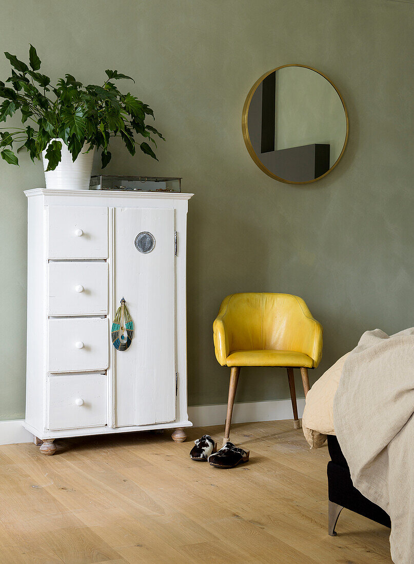 White wardrobe with plant, yellow chair and round mirror on green wall in bedroom